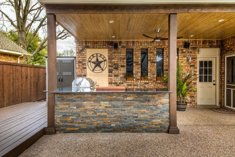 Covered backyard patio with outdoor kitchen, brick wall, wooden deck, and potted palm plant